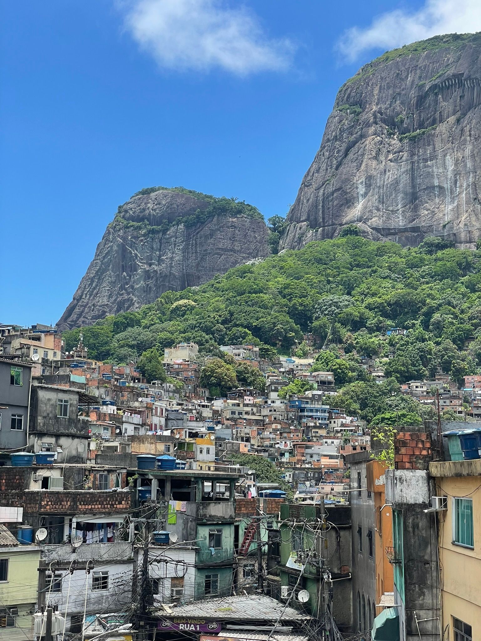 Un'ampia veduta di edifici colorati di una favela, ammassati su una verde collina di fronte a ripide montagne rocciose, sotto un cielo azzurro.