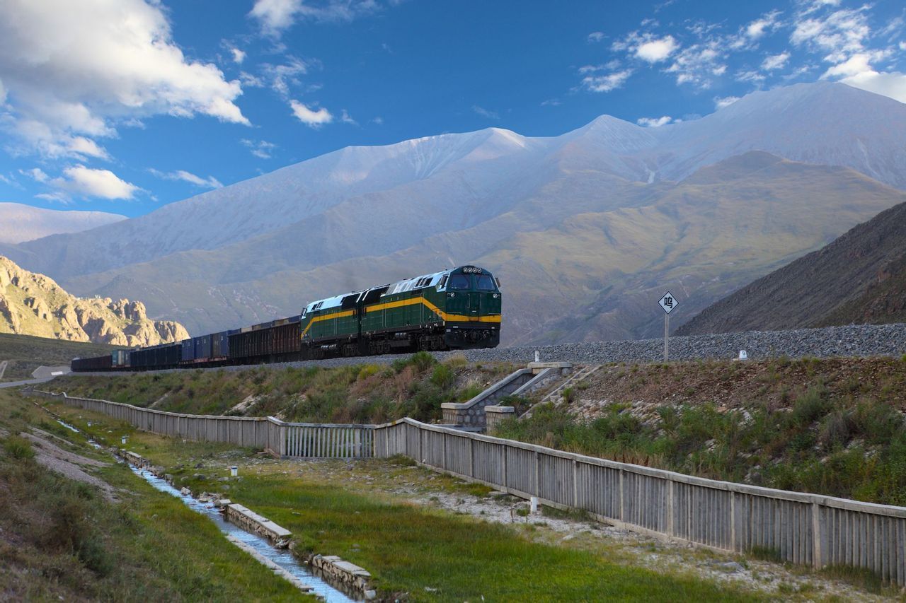Un treno merci verde e giallo viaggia su un binario attraverso una vasta valle di montagna sotto un cielo blu con nuvole bianche.