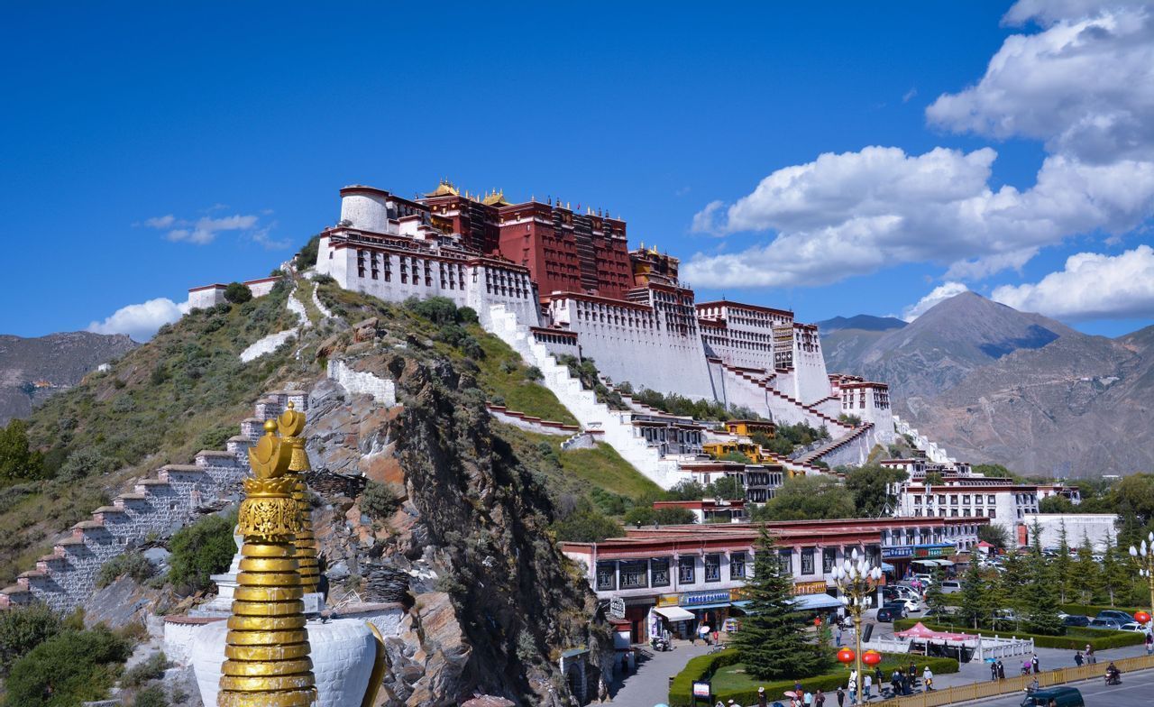 Un grande edificio bianco e rosso su una collina sotto un cielo blu, con uno stupa dorato in primo piano e montagne sullo sfondo.