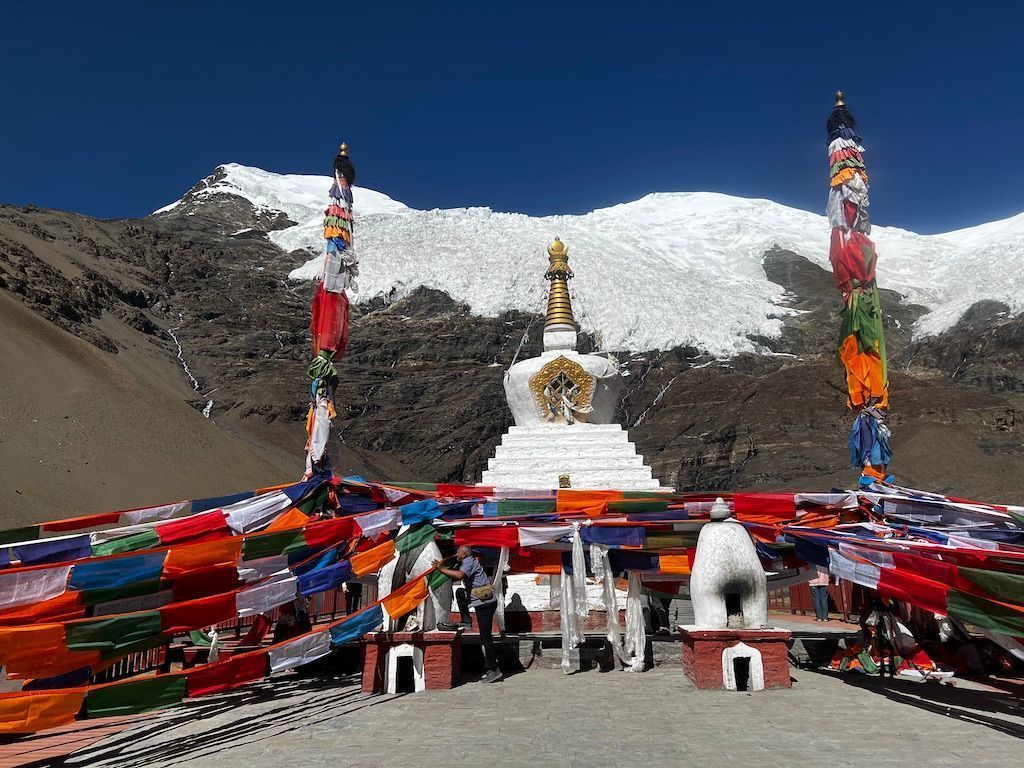 Un bianco stupa buddista è adornato con bandiere di preghiera colorate, con una grande montagna innevata sullo sfondo sotto un cielo sereno.
