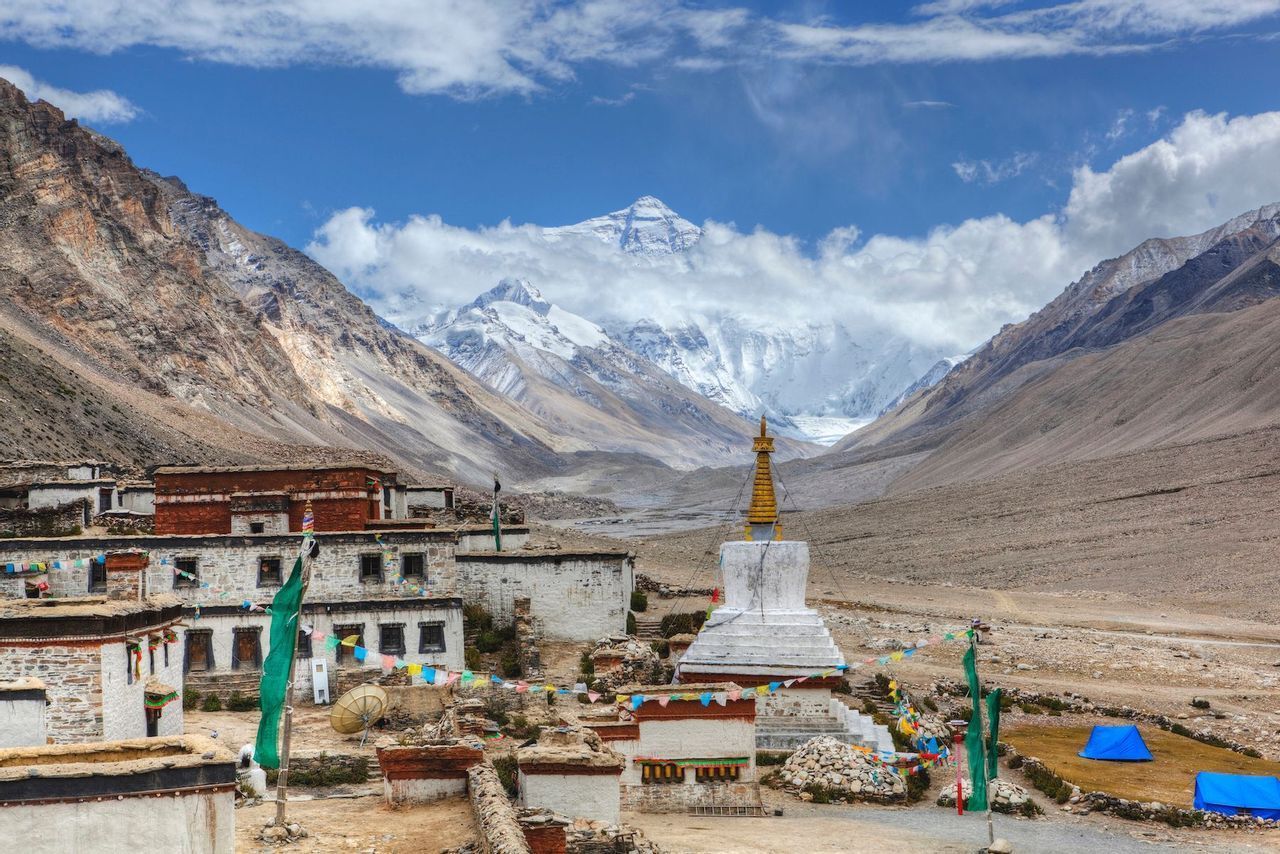 Un villaggio ad alta quota con uno stupa bianco e bandiere di preghiera si trova in una valle di fronte a un'imponente montagna innevata.