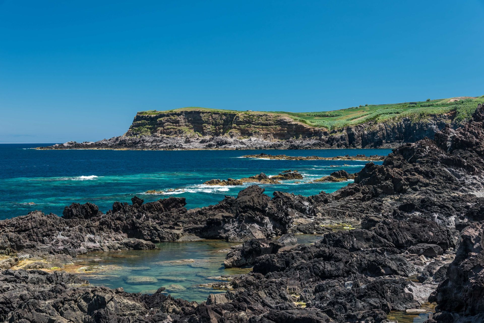 Un littoral rocheux et volcanique avec des piscines naturelles, une mer turquoise et des falaises vertes lointaines sous un ciel bleu clair.