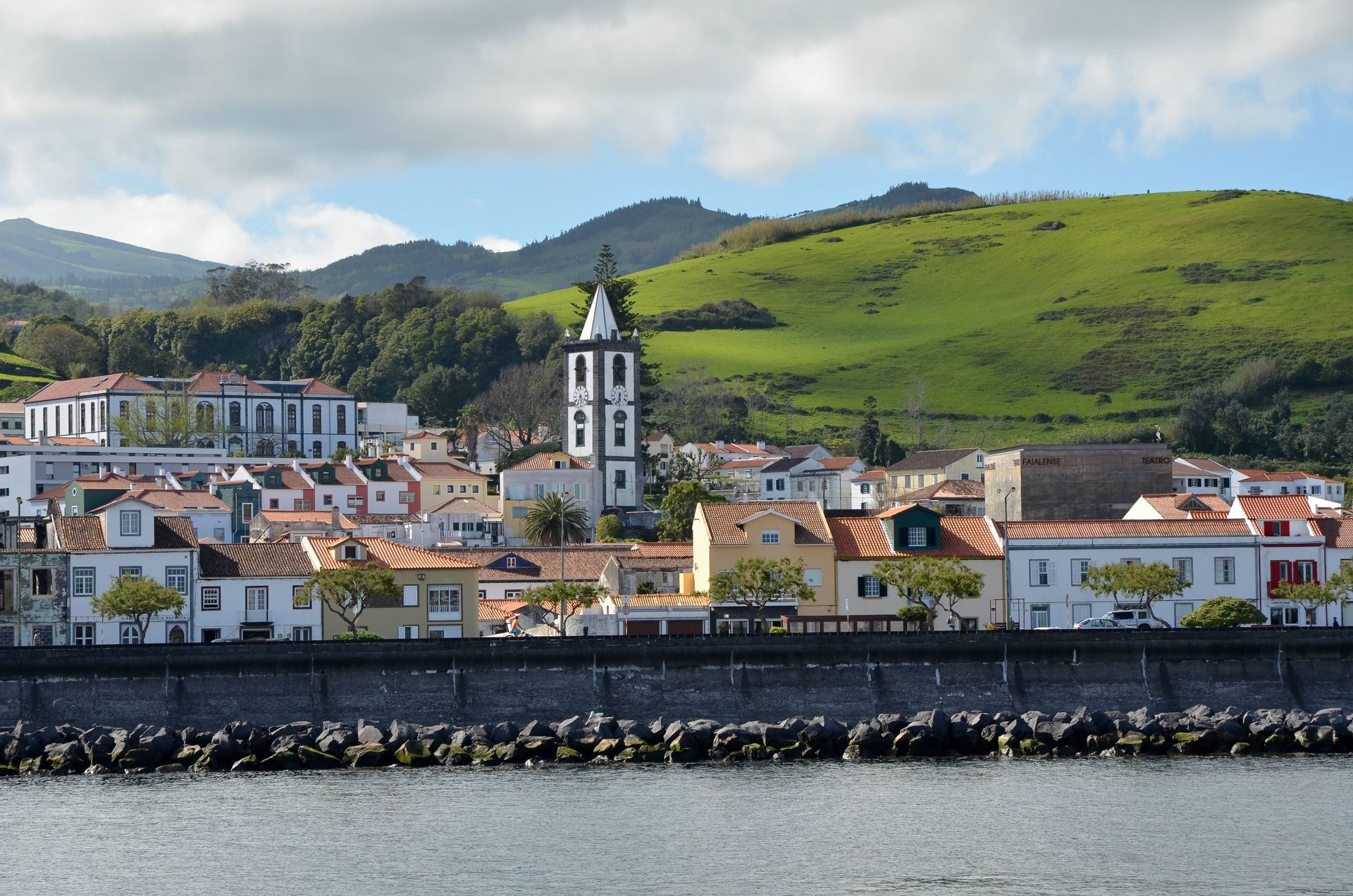 Une ville côtière avec des bâtiments colorés et un clocher d'église sombre se dresse derrière une digue en pierre, avec une grande colline verte en arrière-plan.