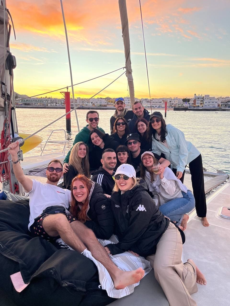 Un gruppo WeRoad sorride per una foto sul ponte di una barca a vela al tramonto, con una città costiera sullo sfondo.
