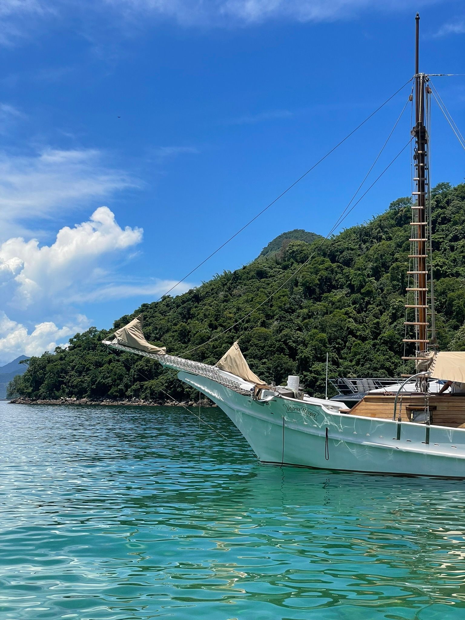 La prua di una barca a vela bianca galleggia su acque tranquille e turchesi vicino a un'isola lussureggiante e alberata sotto un cielo azzurro.