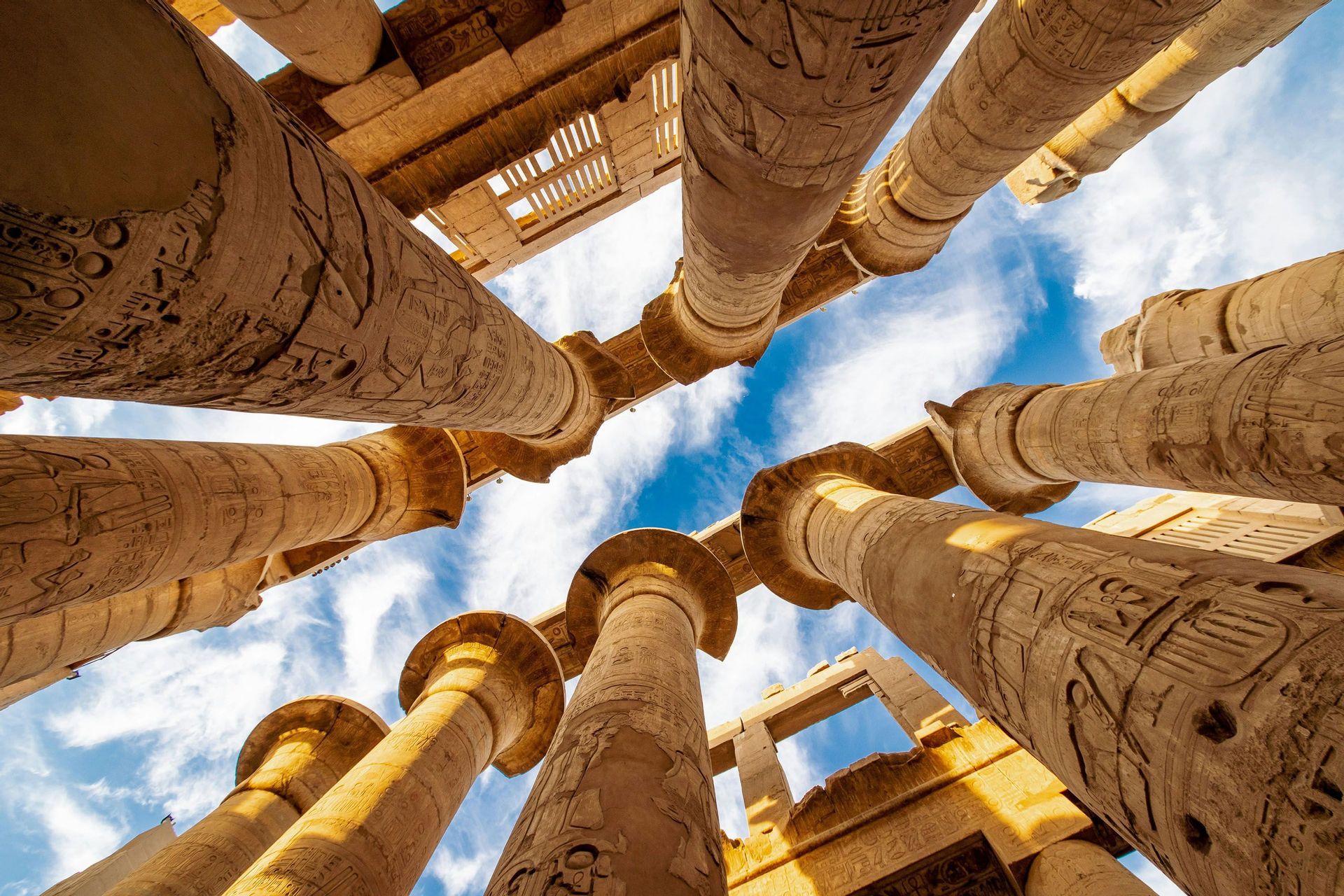 Una vista en contrapicado de grandes columnas de piedra con jeroglíficos antiguos que se elevan hacia un cielo azul y nublado.