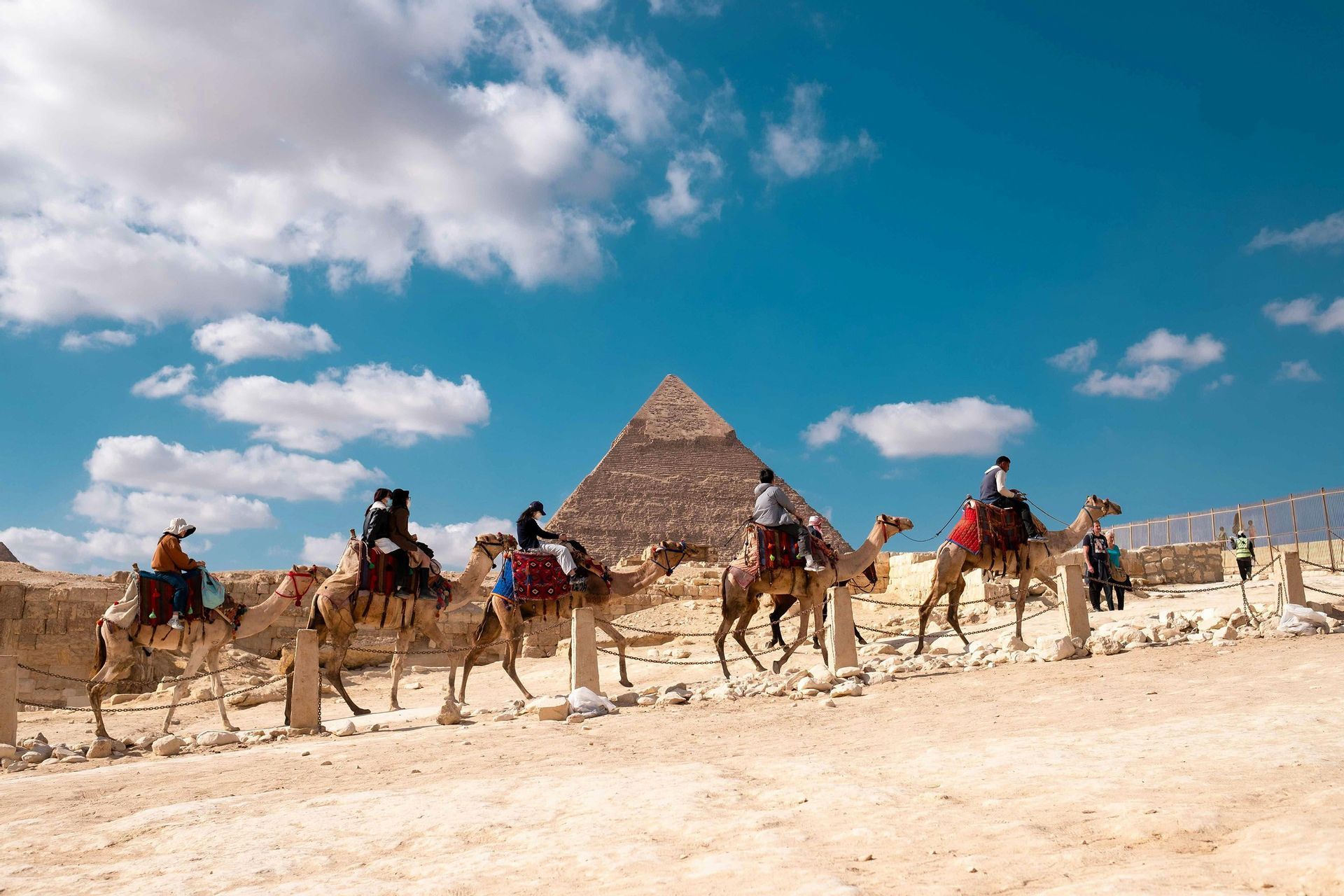 Un viaje en grupo de WeRoad montando en una fila de camellos en el desierto, con una gran pirámide al fondo bajo un cielo azul con nubes.