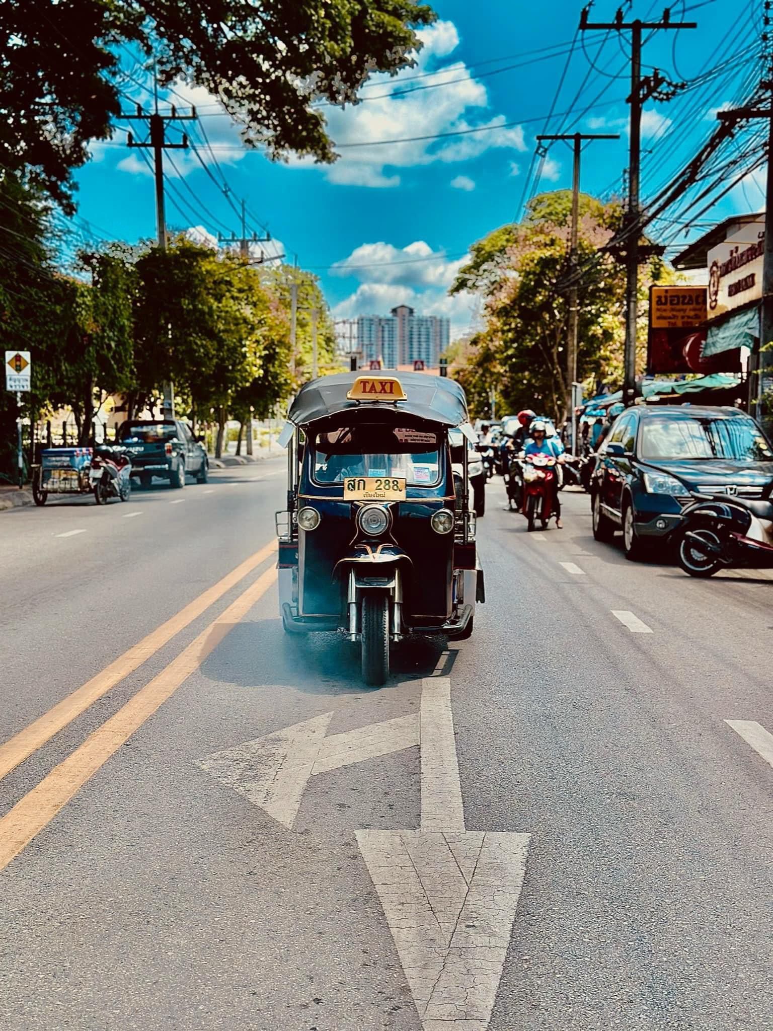 Un taxi tuk-tuk azul circula por una concurrida calle de la ciudad, pasando sobre una gran flecha pintada, con árboles y cables eléctricos en lo alto.