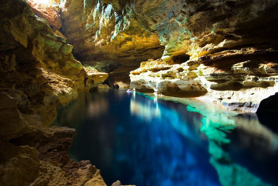 An underground cave with a natural pool of vibrant blue water, illuminated by a beam of sunlight from an opening above.
