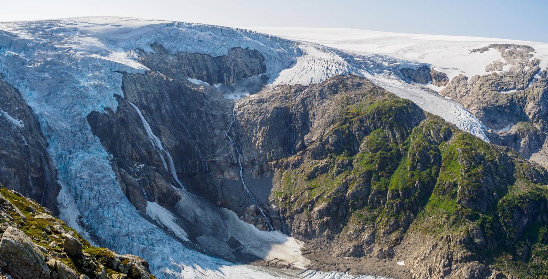 Ein großer Gletscher auf einem felsigen Berg, wobei Schmelzwasser Wasserfälle bildet, die seine steilen Hänge hinabfließen.