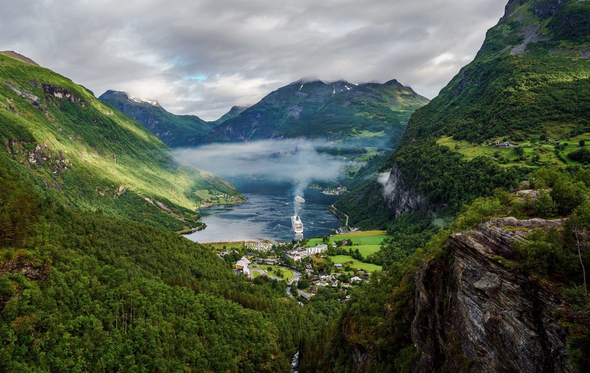 Erhöhter Blick auf zwei Kreuzfahrtschiffe, die durch einen Fjord gleiten, umgeben von steilen, bewaldeten Bergen und einer kleinen Stadt.