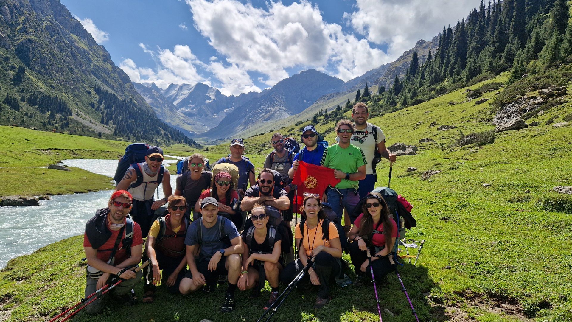 Un groupe WeRoad en voyage avec sacs à dos et bâtons de trekking pose pour une photo dans une verte vallée de montagne, l'un d'eux brandissant un drapeau du Kirghizistan.