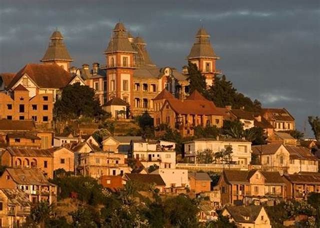 A dense cluster of historic buildings with towers sits on a hillside, illuminated by the warm light of sunset against a dark sky.