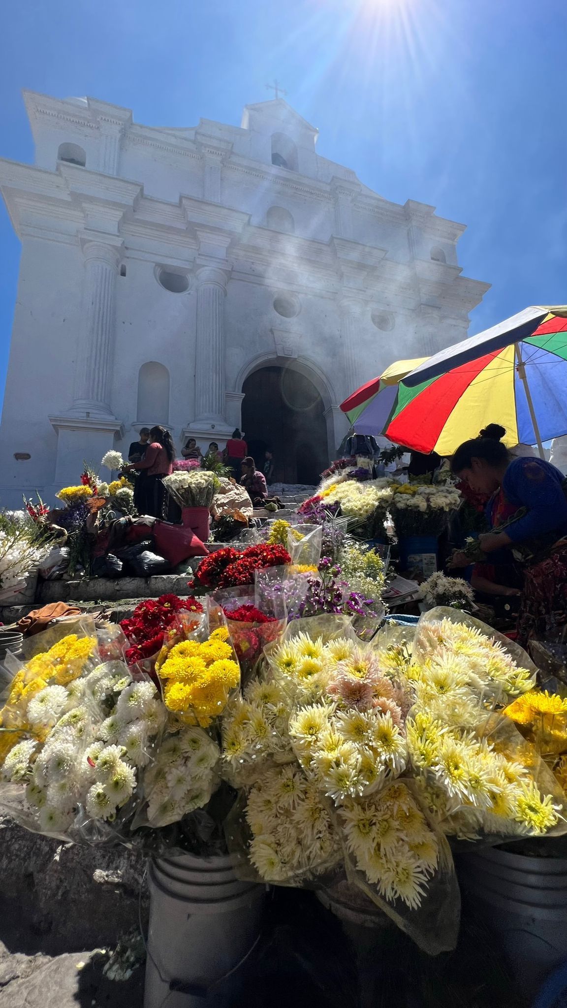 An outdoor flower market with colorful bouquets displayed on the steps of a large, white colonial church under a bright sun.