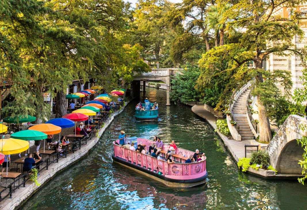 Due battelli turistici con passeggeri scivolano lungo un canale costeggiato da caffè all'aperto, protetti da ombrelloni colorati e alberi rigogliosi.