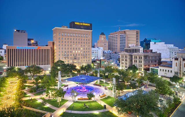 Un parco cittadino illuminato con una fontana centrale e vialetti al crepuscolo, con uno skyline di edifici moderni sullo sfondo.