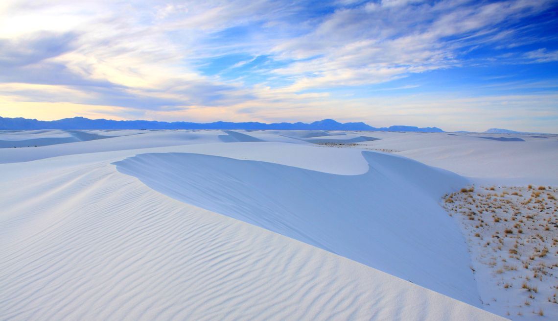 Dune di sabbia bianche ondulate con increspature del vento si estendono verso montagne lontane sotto un cielo parzialmente nuvoloso.