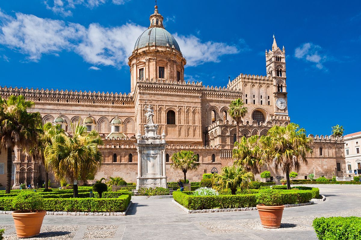 Una gran catedral histórica con cúpula se erige detrás de un cuidado jardín con palmeras bajo un cielo azul.