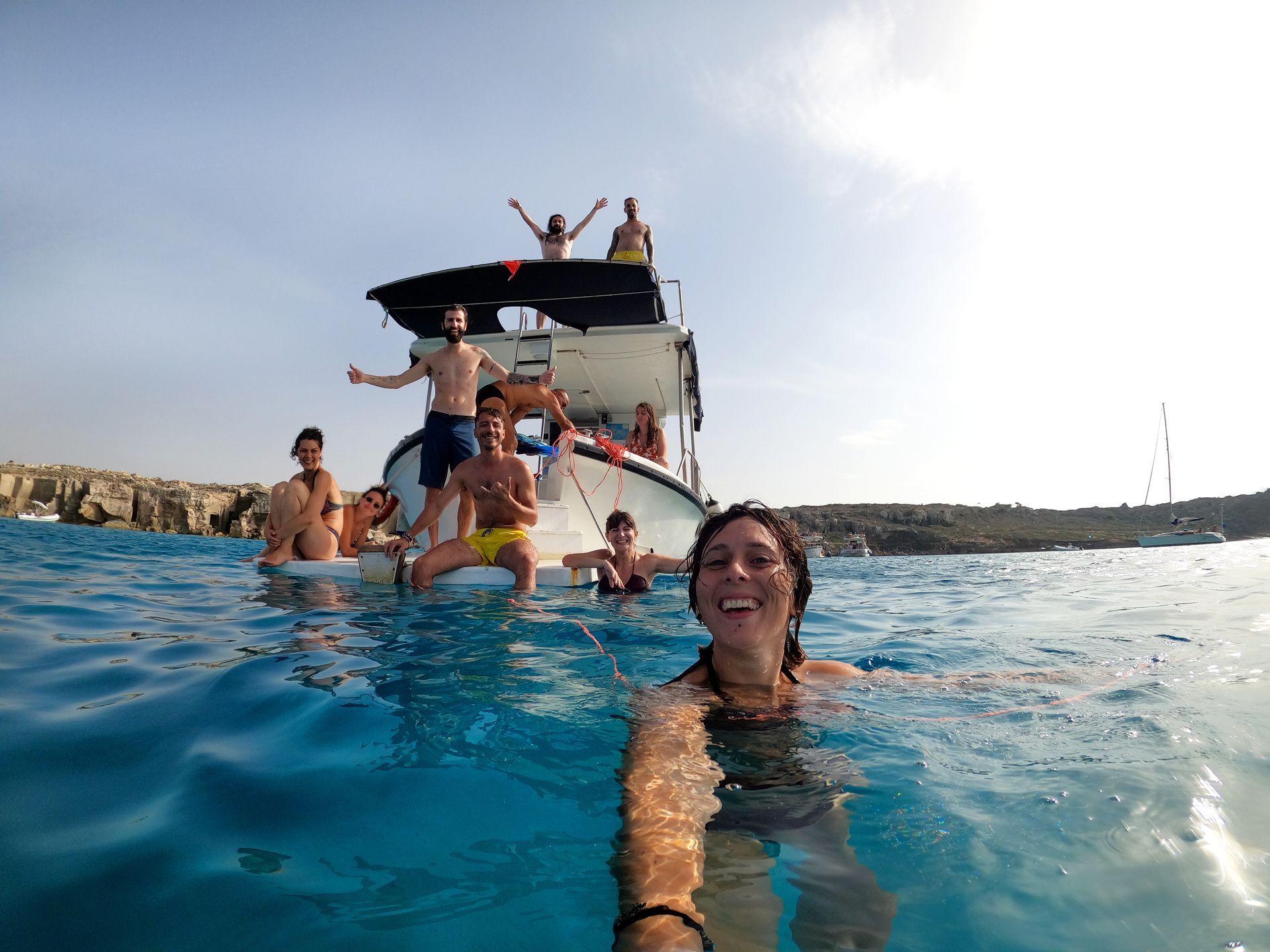 Una mujer se toma una selfie sonriendo mientras nada en el mar con su grupo de viaje WeRoad relajándose en un barco al fondo.