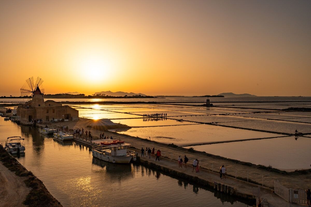 Un viaje en grupo de WeRoad observa el atardecer dorado sobre las salinas junto a un canal con barcos amarrados y un molino de viento tradicional.