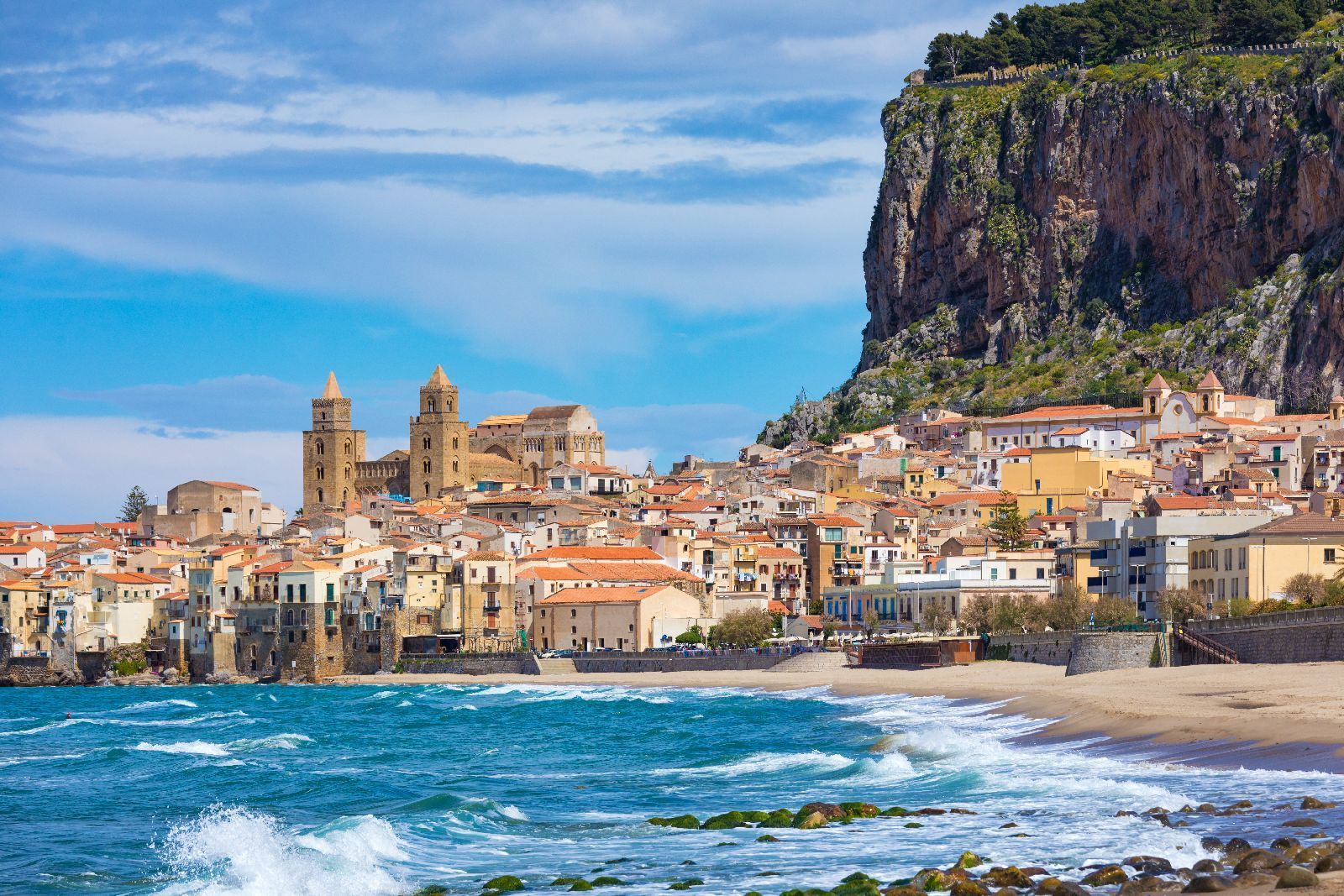 Un pueblo costero con una catedral, enclavado al pie de un gran acantilado, junto a una playa de arena con olas.