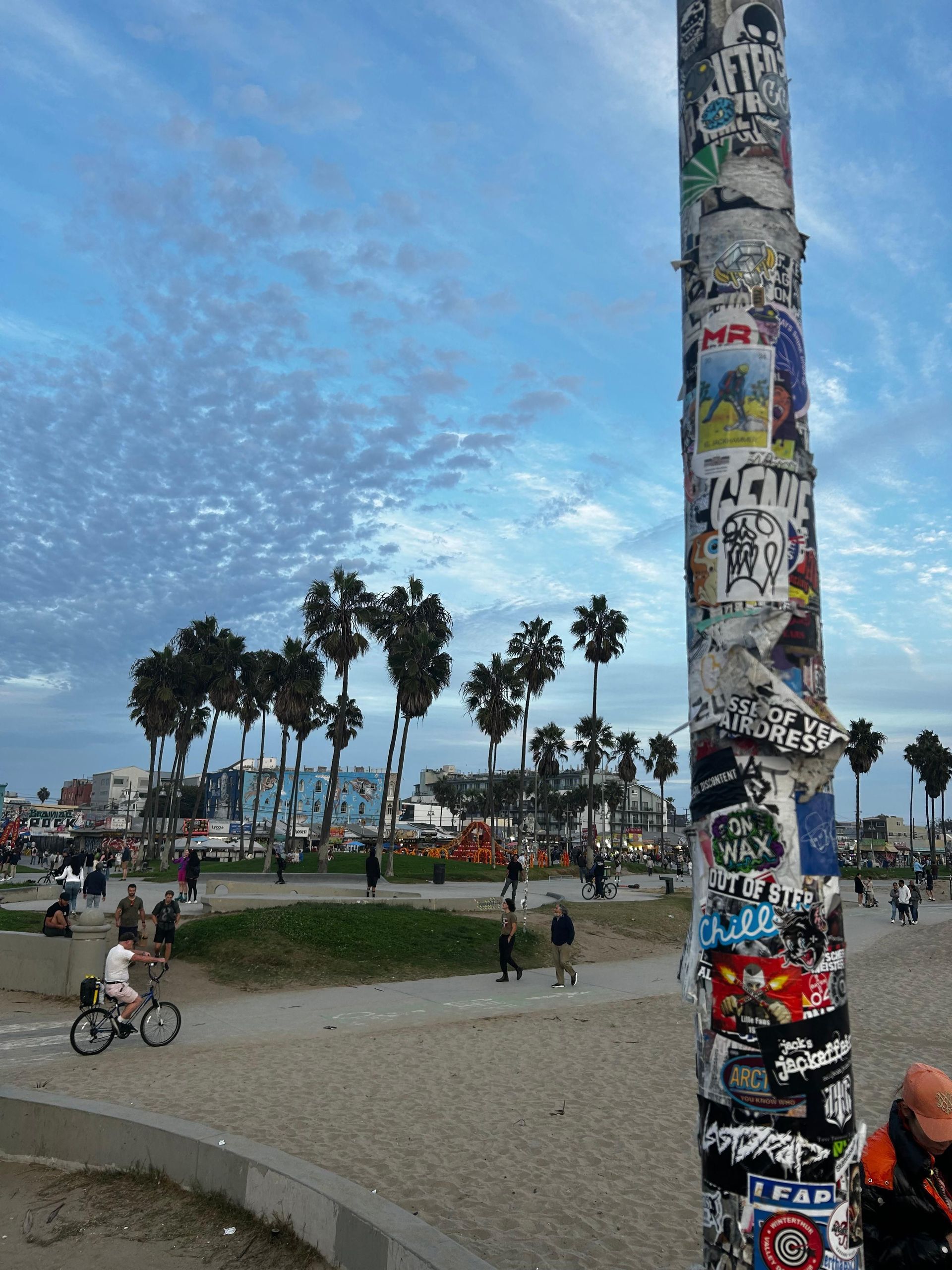 Un poste cubierto de pegatinas se alza en una playa, con gente caminando y palmeras bajo un cielo nublado al fondo.