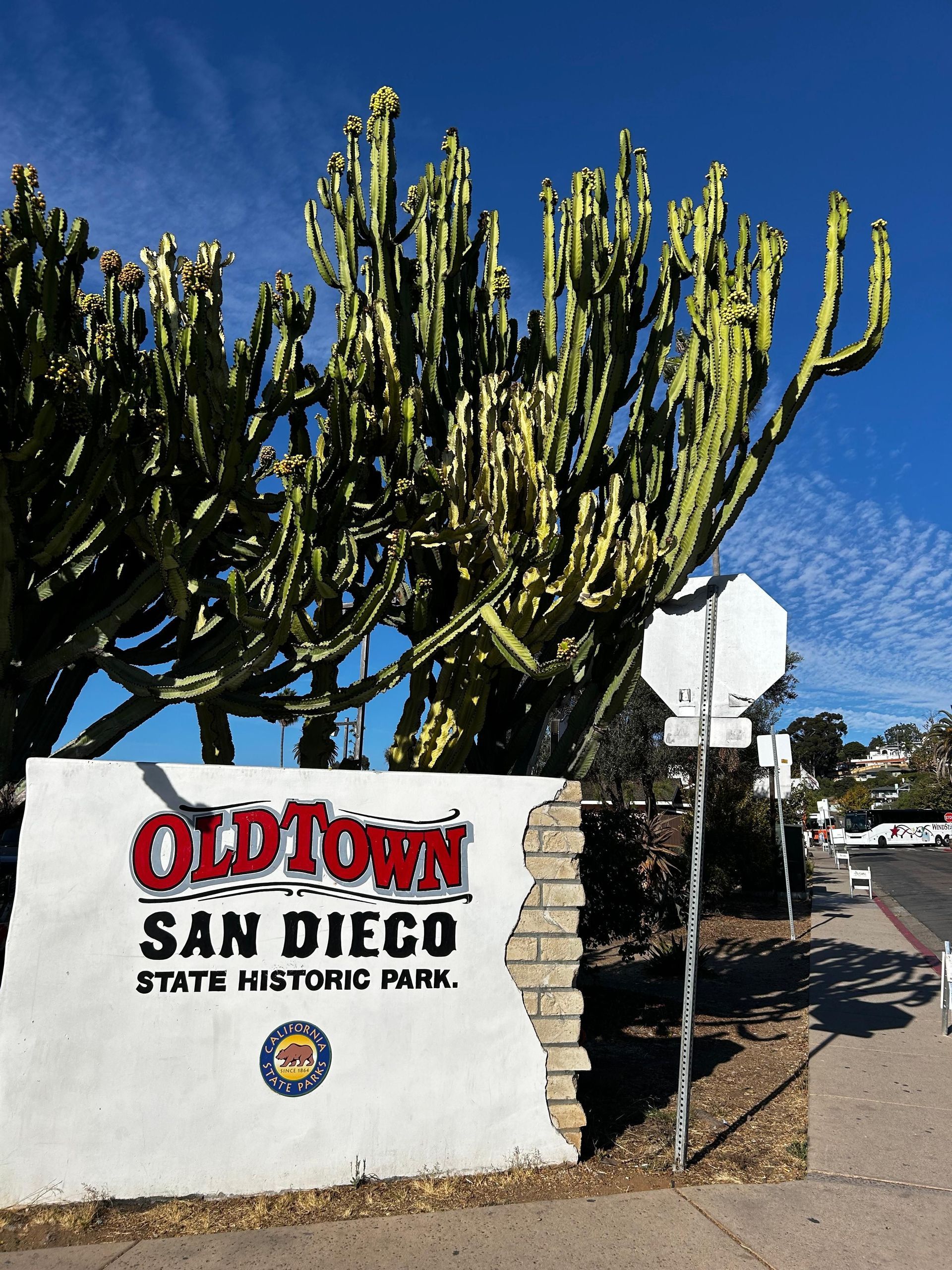 El letrero blanco de entrada del Parque Histórico Estatal Old Town San Diego se alza frente a un gran cactus bajo un cielo azul.
