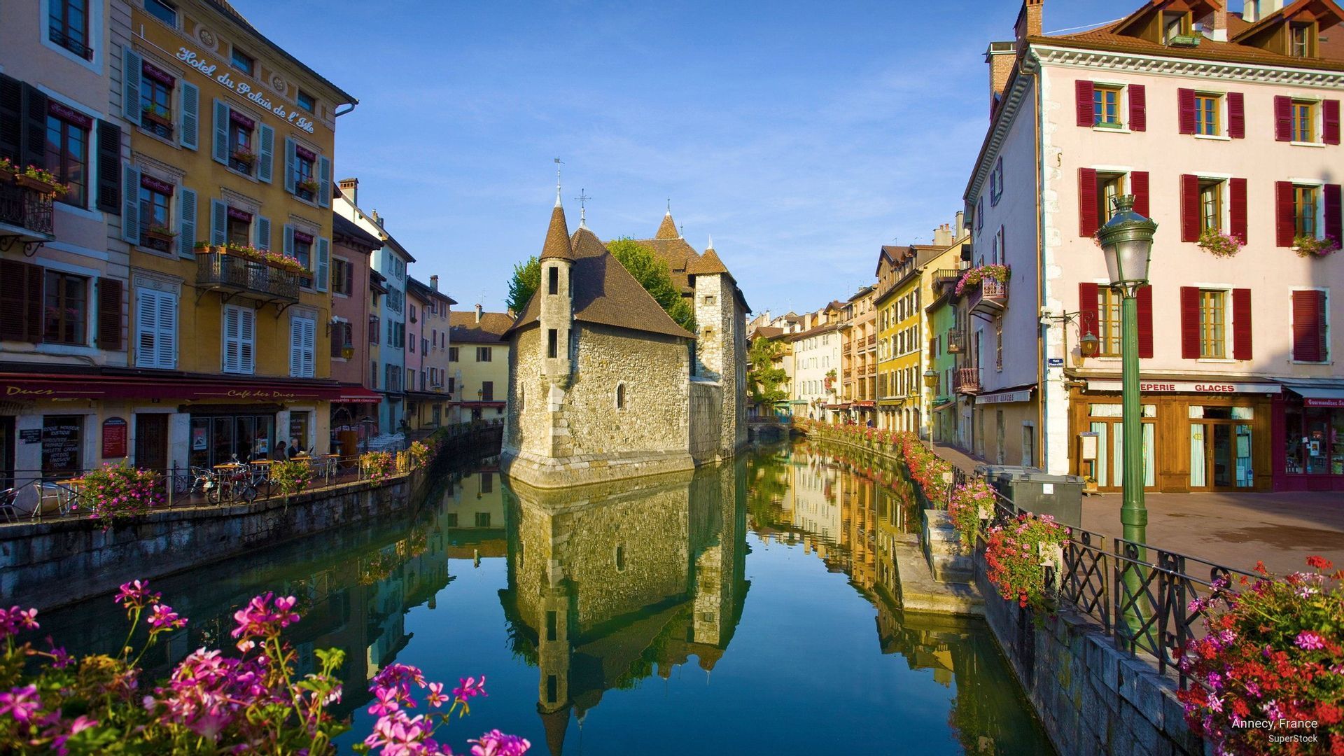 Un edificio storico in pietra si erge su un canale, fiancheggiato da edifici colorati le cui riflessioni sono visibili nell'acqua calma.