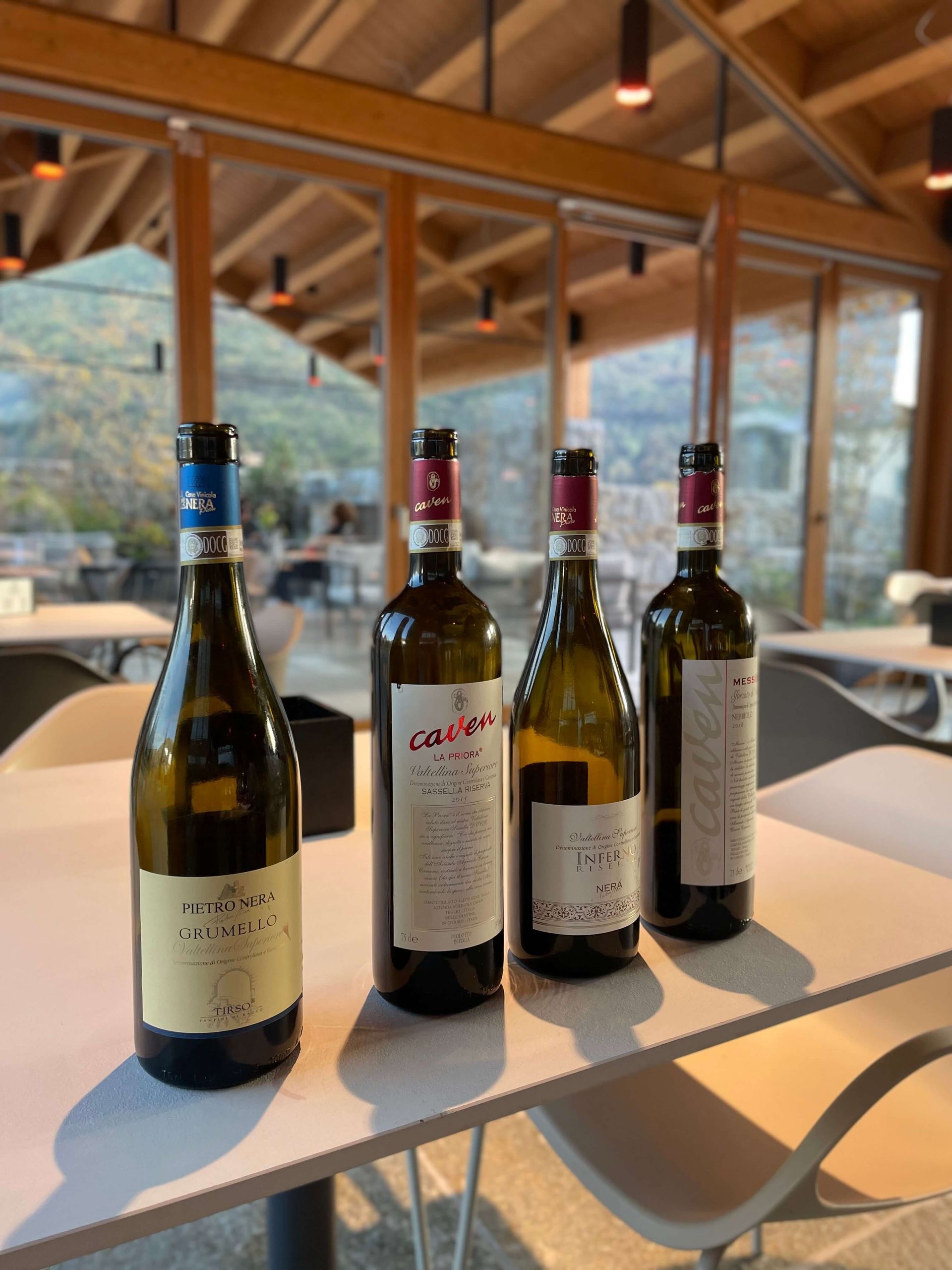 Quattro bottiglie di vino sono disposte su un tavolo bianco per una degustazione, in una sala moderna con ampie finestre che si affacciano su una collina.