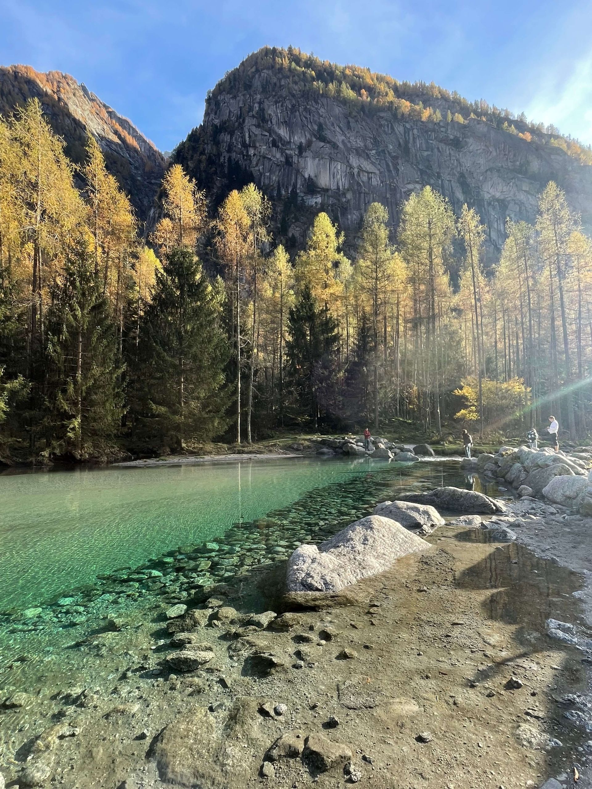 Un viaggio di gruppo WeRoad sulla riva di un lago limpido e turchese, ai piedi di una montagna soleggiata e di una foresta autunnale.