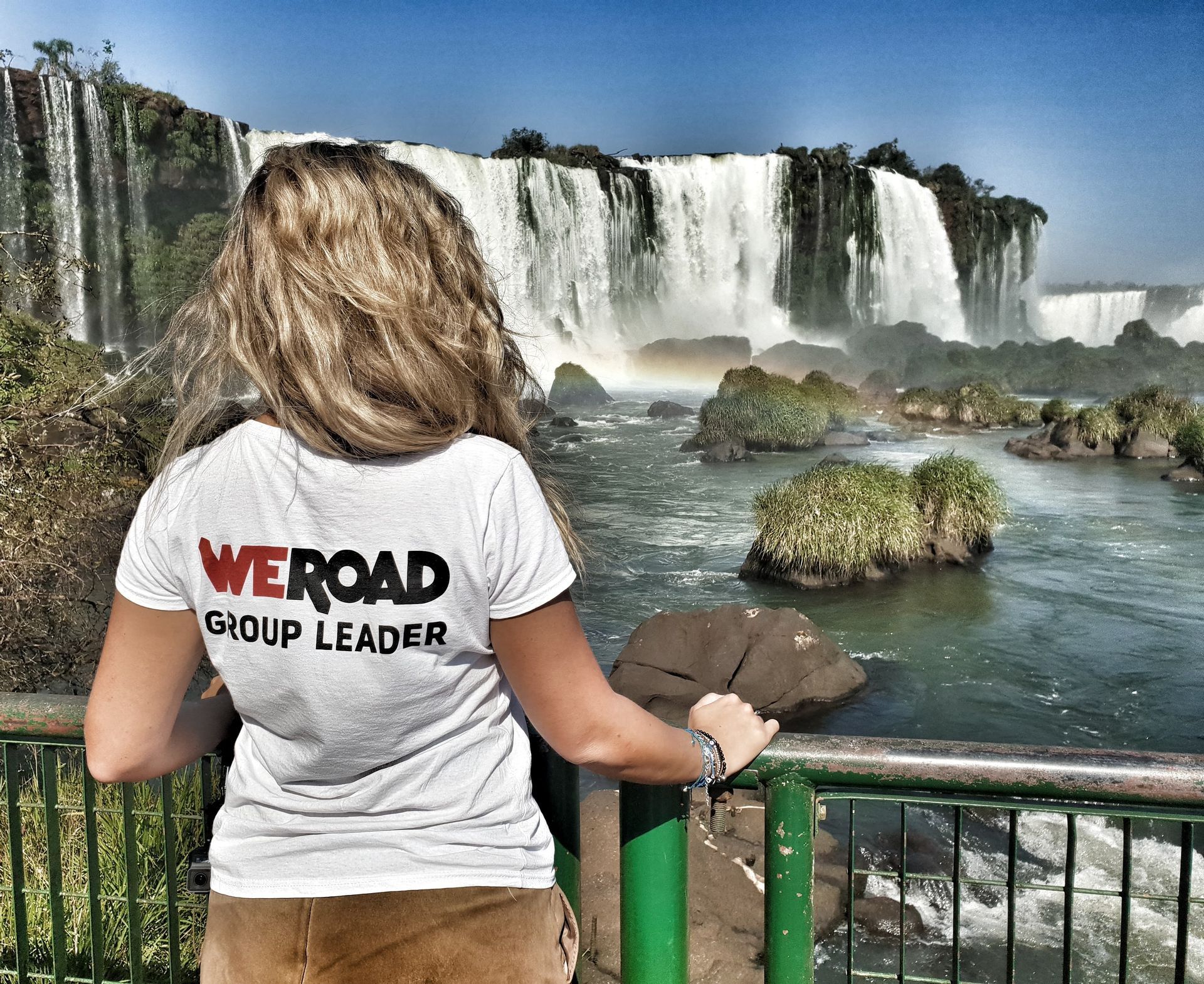 A WeRoad group leader with blonde hair stands at a railing, looking out at a large, cascading waterfall under a clear blue sky.