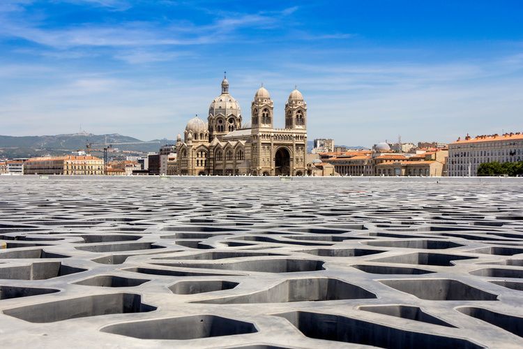 Una vista de una gran catedral a través de una moderna terraza de hormigón estampado, con un paisaje urbano de fondo bajo un cielo azul.