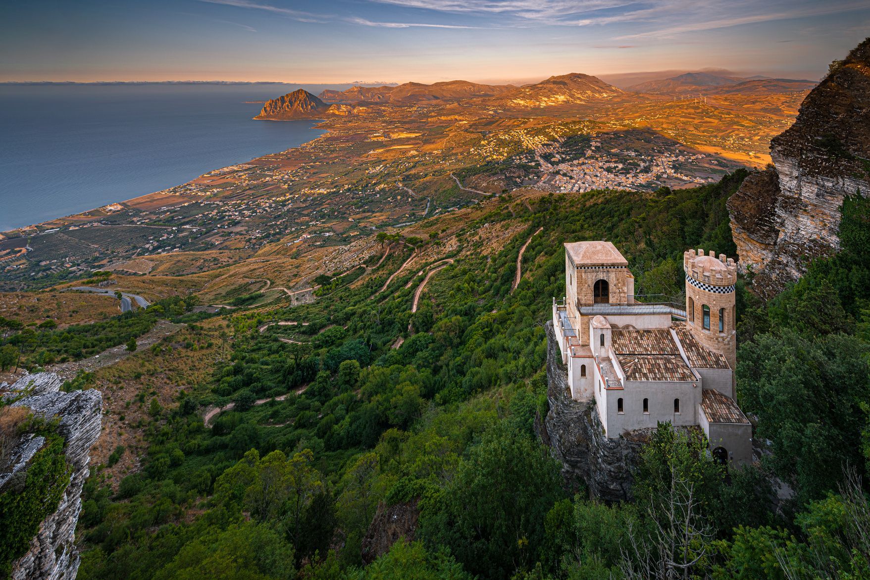 Un castello di pietra con una torre si erge su una scogliera che domina una vasta costa e montagne al tramonto.