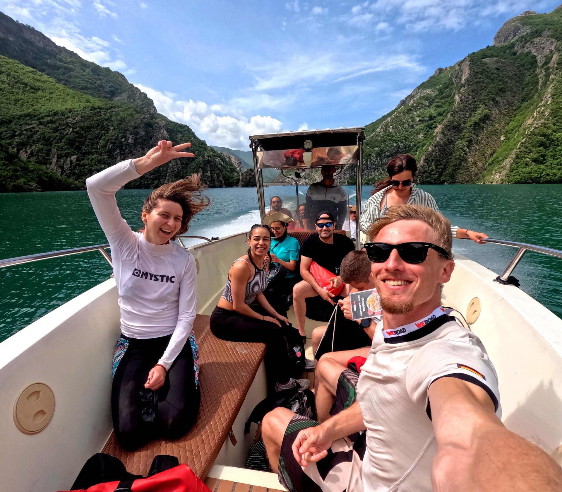Un grupo de WeRoad se hace una selfie en un barco, rodeado de empinadas montañas verdes y agua azul.