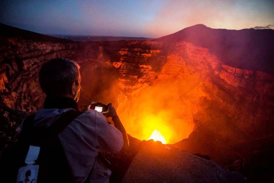 Une personne avec un sac à dos photographie un cratère de lave incandescent à l'intérieur d'un volcan au crépuscule.