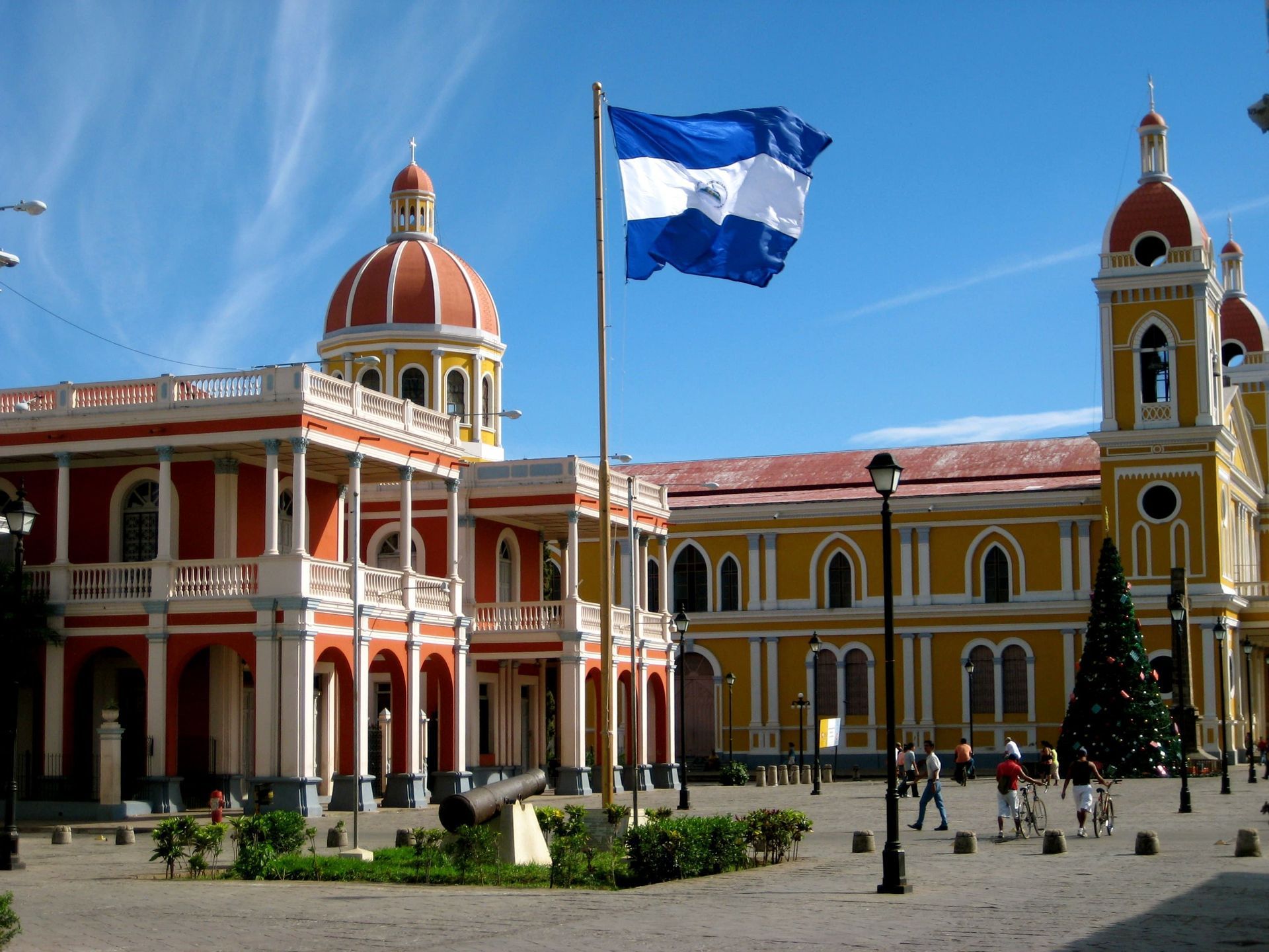 Le drapeau du Nicaragua flotte au-dessus d'une place coloniale ensoleillée avec des bâtiments rouges et jaunes, des dômes et un canon.