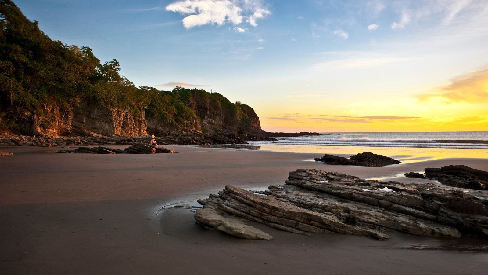 Une personne assise sur un rocher sur une plage de sable, avec des falaises couvertes d'arbres et l'océan visibles lors d'un coucher de soleil doré.