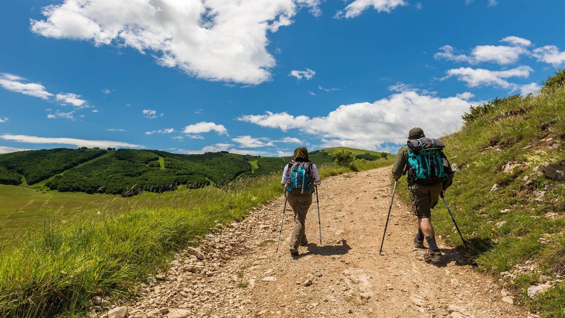 Due persone con zaini e bastoncini da trekking camminano su un sentiero sterrato su una montagna verde sotto un cielo azzurro con nuvole.