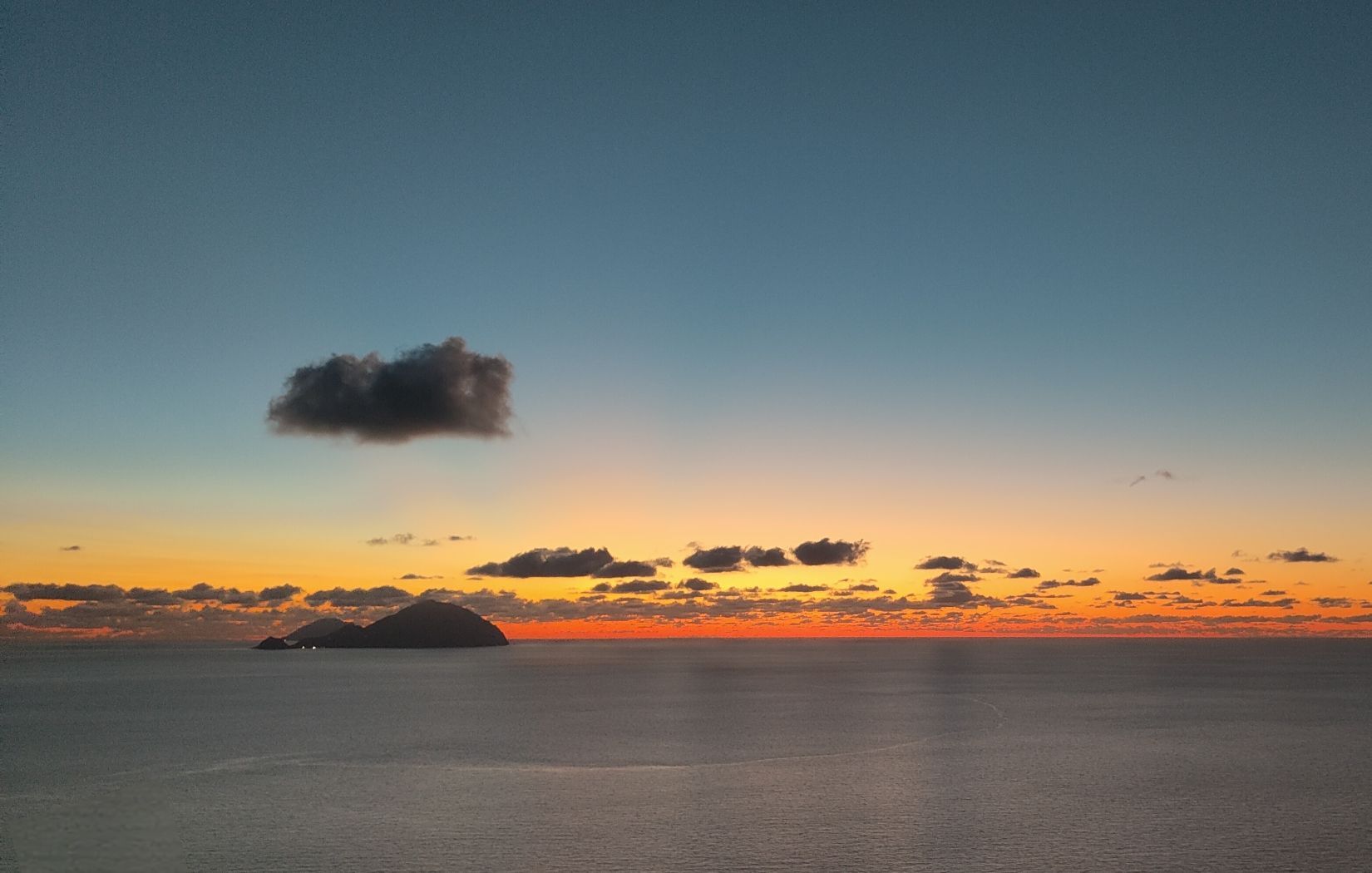 A sunset colors the sky orange and blue over a calm ocean, with a silhouetted island on the horizon.