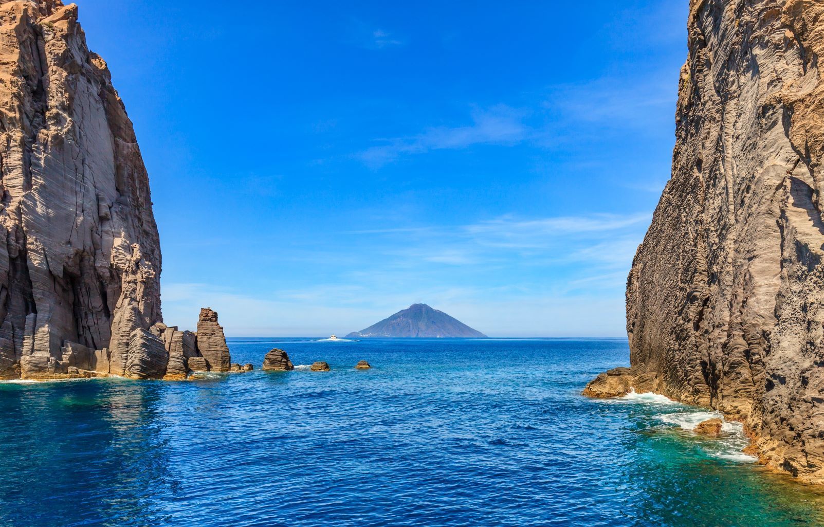 A distant volcanic island is viewed from the sea between two large, rocky cliffs under a clear blue sky.