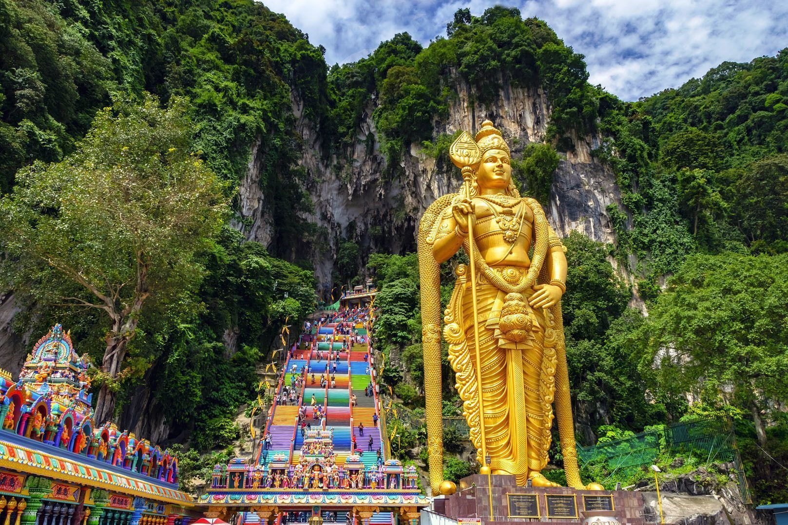 Una gigantesca statua dorata si erge accanto a una lunga scalinata dai colori dell'arcobaleno che porta su una rigogliosa scogliera verde, mentre le persone la salgono.