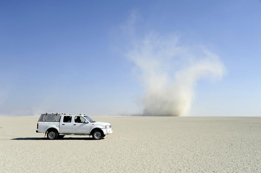 Un pick-up bianco è parcheggiato su una vasta, piatta pianura desertica mentre un grande diavolo di sabbia vortica in lontananza sotto un cielo azzurro e limpido.
