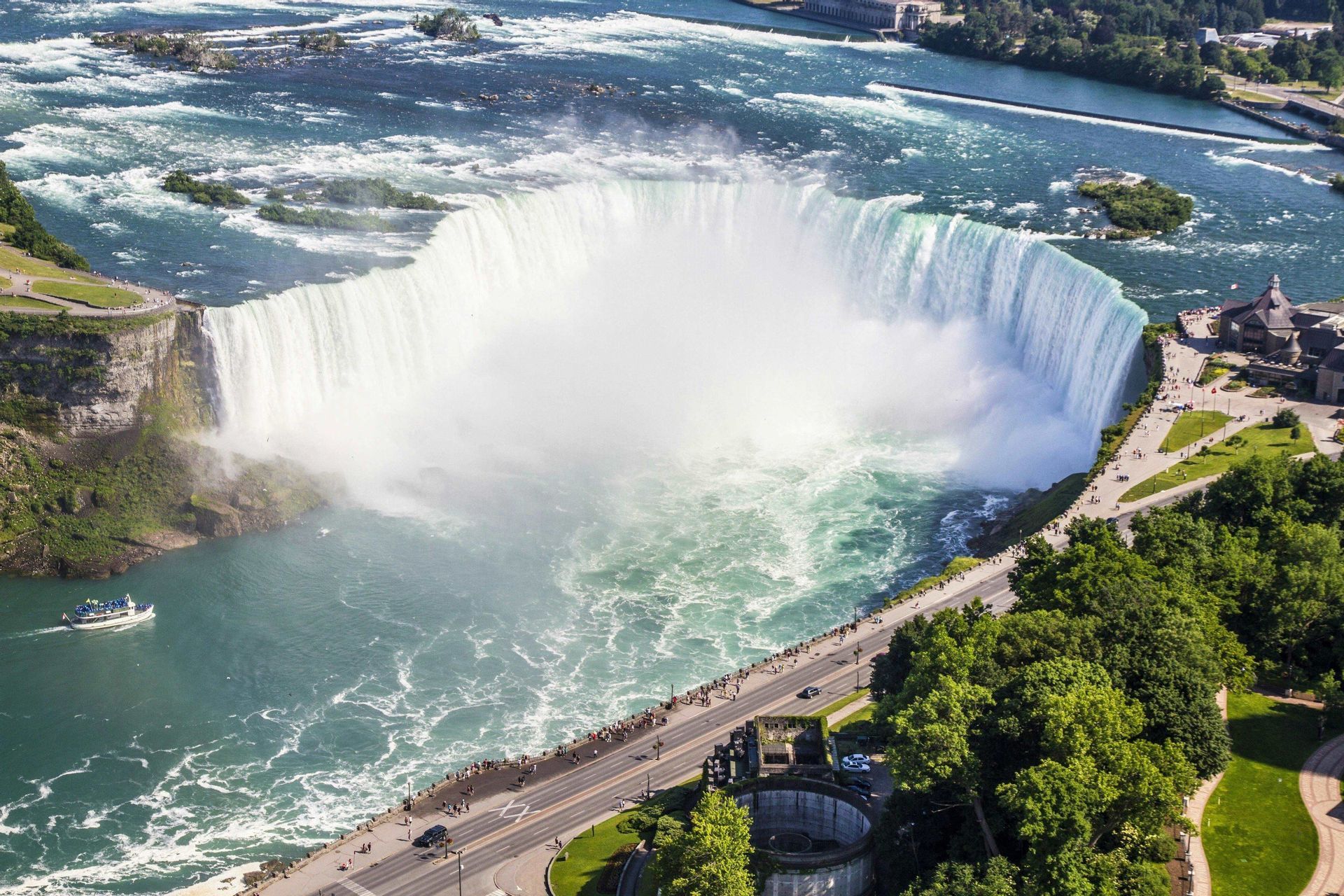 Una vista aerea di una massiccia cascata a forma di ferro di cavallo che si getta in un fiume, con una barca turistica sotto e persone su una passeggiata vicina.