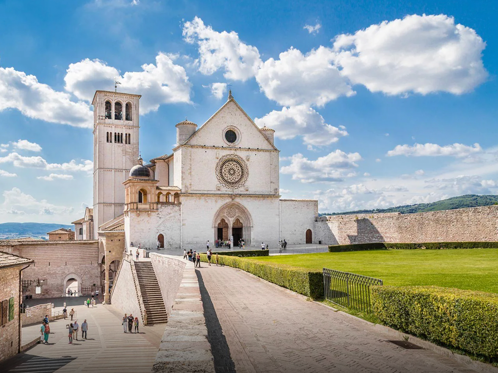 Una grande basilica in pietra bianca con un campanile e un rosone si erge sotto un cielo blu, con un sentiero lastricato che conduce al suo ingresso.
