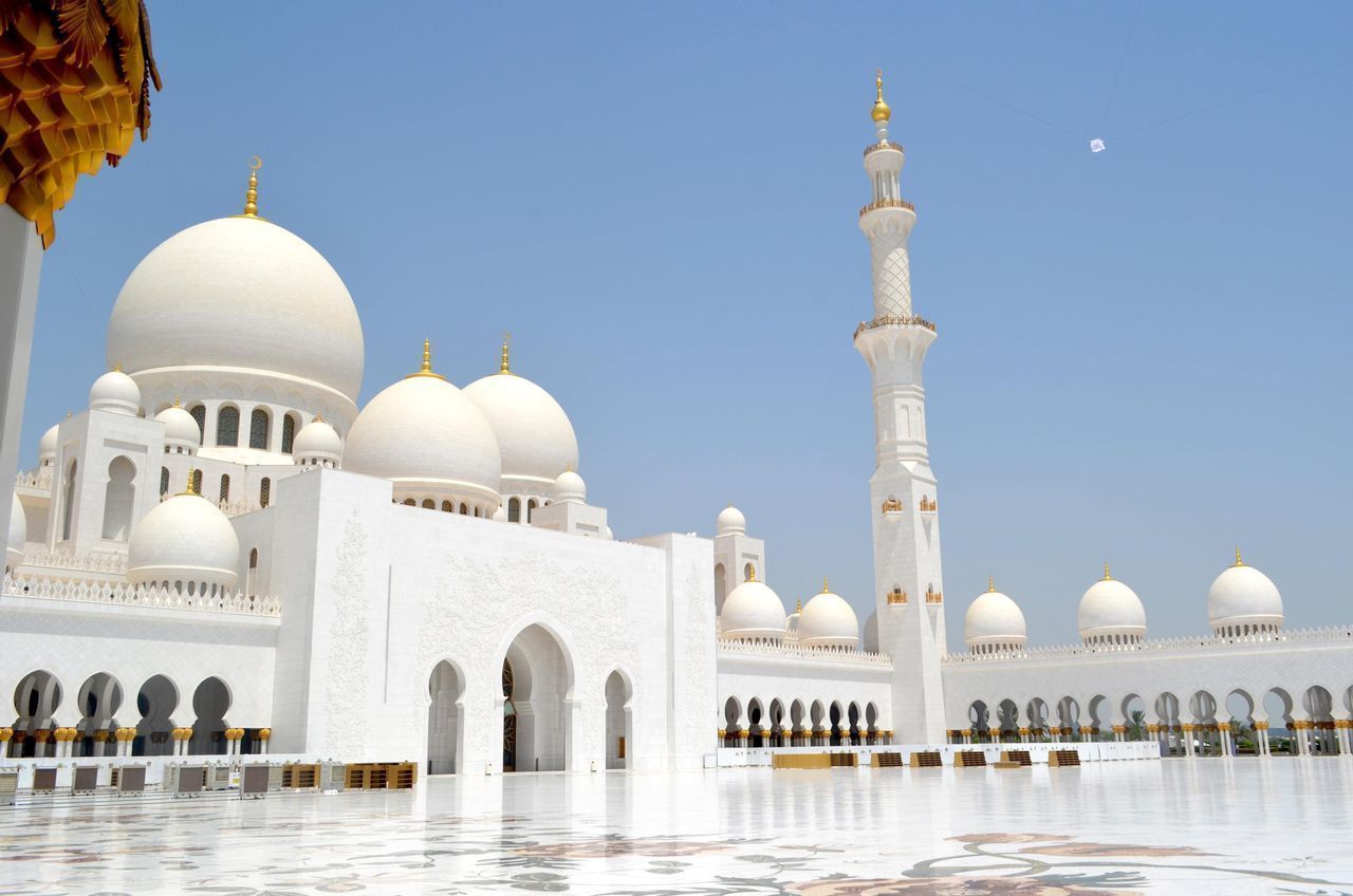 Una grande moschea bianca con cupole multiple e un minareto, vista da un cortile di marmo riflettente sotto un cielo azzurro limpido.