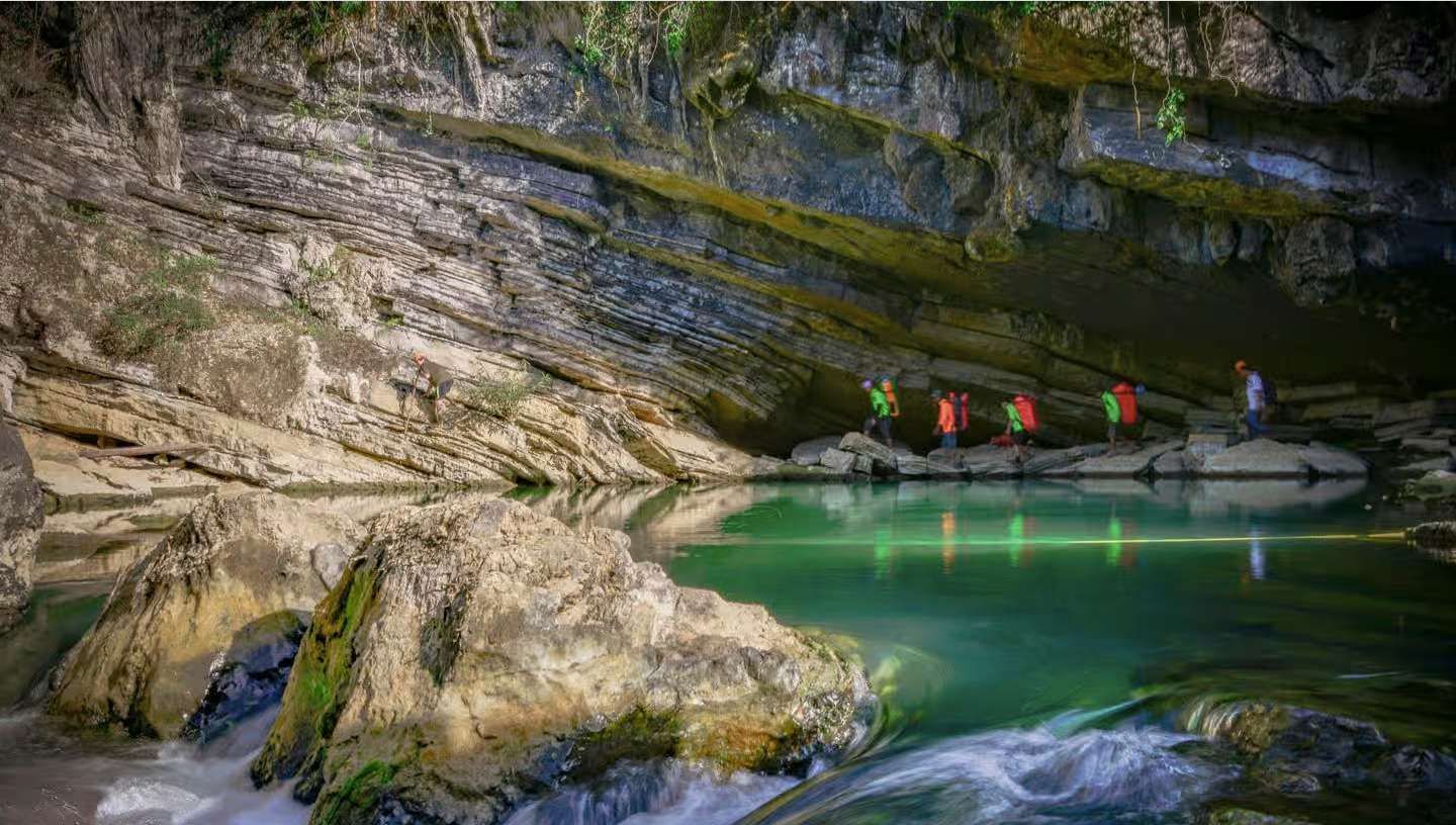Un viaggio di gruppo WeRoad esplora un'ampia grotta, camminando lungo un sentiero roccioso accanto a un fiume verde smeraldo.