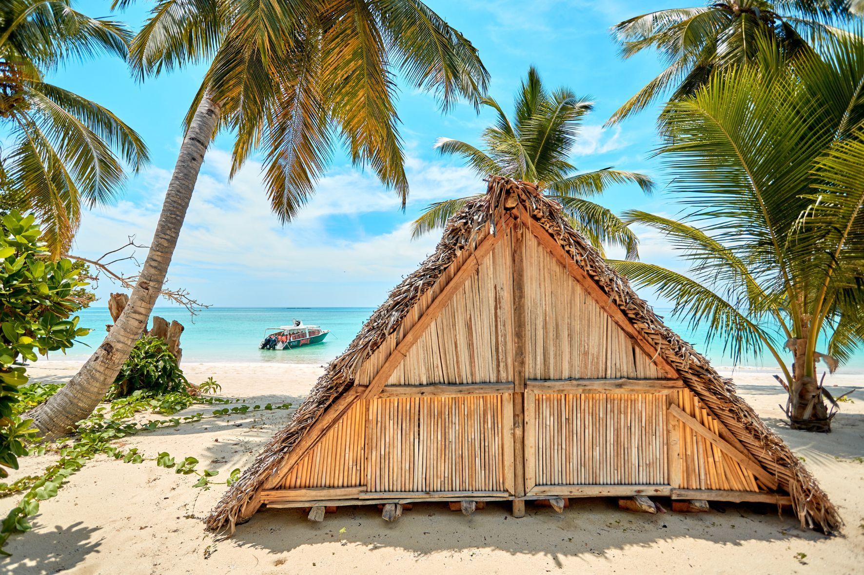 Eine dreieckige Holzhütte mit Strohdach steht an einem weißen Sandstrand unter Palmen, mit einem Boot auf dem türkisfarbenen Wasser.