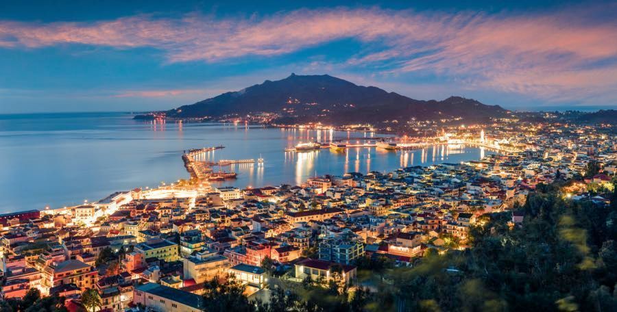 Una vista aérea de una ciudad costera y su puerto iluminados al anochecer, con una gran montaña elevándose detrás del agua.