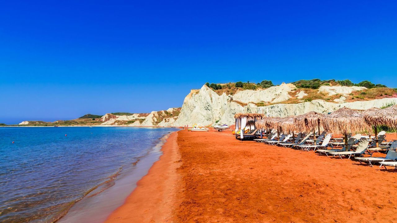 Tumbonas y sombrillas de paja se alinean en una playa de arena naranja junto a aguas azules tranquilas, con acantilados blancos bajo un cielo despejado.