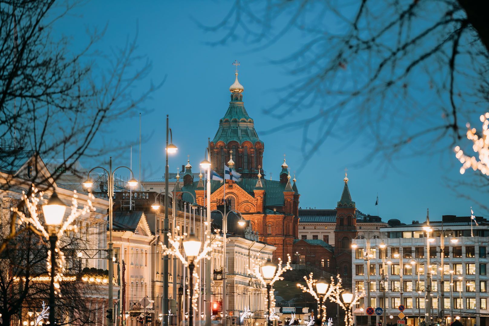 Una via della città decorata con luci festive porta verso una grande cattedrale in mattoni rossi con cupole verdi al crepuscolo.